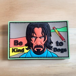 May include: Rectangular patch with a green border featuring a stylized image of a man with black hair and a beard. The text "Be Kind to Dogs" is displayed with a pencil graphic.