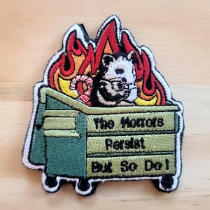 May include: Embroidered patch featuring a raccoon in a dumpster engulfed in flames. The patch has a green dumpster with the text "The Horrors Persist But So Do I" in white. The flames are red, orange, and yellow.