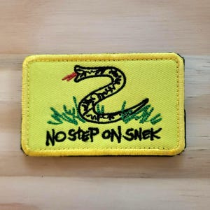 May include: Embroidered patch featuring a coiled black snake with a red tongue on a yellow background. The patch has the text "NO STEP ON SNEK" in black. The patch is rectangular with a yellow border.