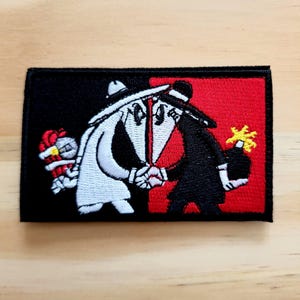May include: Embroidered patch featuring two cartoon figures shaking hands, one white and one black, on a black and red background. The white figure holds a bouquet, and the black figure holds a bomb. The patch is rectangular.