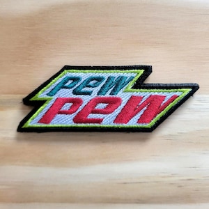 May include: Embroidered patch with the text "PEW PEW" in a stylized design. The top text is teal and the bottom text is red, both outlined in white. The patch has a black border with a green outline.