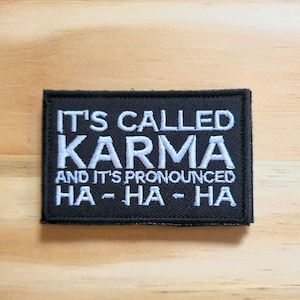 May include: Black rectangular patch with white embroidered text that reads "IT'S CALLED KARMA AND IT'S PRONOUNCED HA - HA - HA". The patch is on a light brown wooden surface.