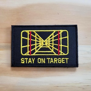 May include: Black rectangular patch with yellow geometric design and red vertical lines. The text "STAY ON TARGET" is embroidered in white. The patch is likely for a jacket or bag.