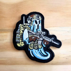 May include: Embroidered patch featuring a ghost soldier in a helmet, tactical vest, and holding a rifle. The ghost is light blue with a black outline, and the patch has a black border. The text "The Tinkering Table" is at the bottom.