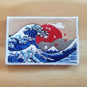 May include: Embroidered patch featuring a stylized depiction of a large ocean wave with a red sun in the background. The wave is rendered in shades of blue and white, with a beige border and a mountain in the background.
