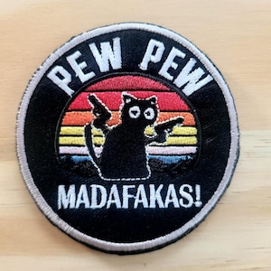 May include: Embroidered patch featuring a black cat silhouette holding two guns, set against a sunset design with the words "PEW PEW MADAFAKAS!" in white lettering on a black background. The patch has a silver border.