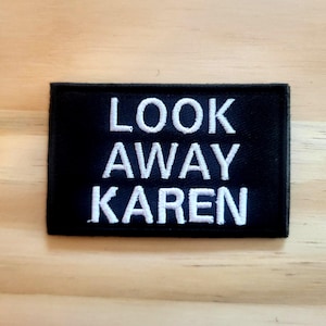 May include: Black rectangular patch with white embroidered text that reads "LOOK AWAY KAREN". The patch is on a light brown wooden surface. The text is in a bold, sans-serif font.