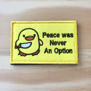 May include: A yellow embroidered patch with a cartoon chick holding a knife. The patch has the text "Peace was Never An Option" in black. The patch is rectangular with a yellow border.