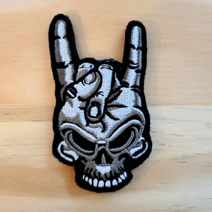 May include: Embroidered patch featuring a skull with the 'horns' hand gesture. The design is in shades of gray and white, with a black border. The patch is likely for clothing or accessories.