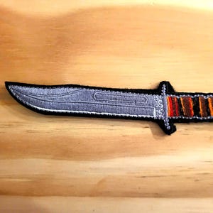 May include: Embroidered patch of a knife with a silver blade and a handle featuring orange, red, and brown stripes. The patch has a black border and is set against a wooden background.