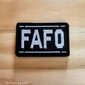 May include: Black rectangular patch with white embroidered text "FAFO". The patch has a white border and is set against a light brown wooden surface. The patch is a rectangular shape.