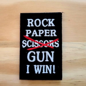 May include: Black rectangular patch with white embroidered text: "ROCK PAPER SCISSORS" with a red "X" over "SCISSORS". Below, the text reads "GUN I WIN!" on a light wood surface.