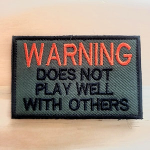 May include: A rectangular patch with the word "WARNING" in orange thread, above the text "DOES NOT PLAY WELL WITH OTHERS" in black thread. The patch has a green background and a black border.