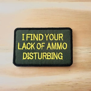 May include: A rectangular, olive green patch with the embroidered text "I FIND YOUR LACK OF AMMO DISTURBING" in yellow. The patch has a black border and is set against a light wood grain background.
