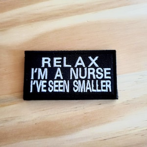 May include: Black rectangular patch with white embroidered text that reads "RELAX I'M A NURSE I'VE SEEN SMALLER". The patch is on a light brown wooden surface.