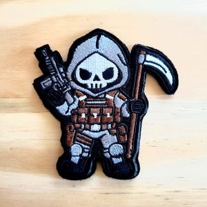 May include: Embroidered patch featuring a cartoon grim reaper character with a skull face, wearing a gray hood and tactical gear. The reaper is holding a rifle in one hand and a scythe in the other. The patch has a black border.