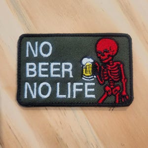 May include: A rectangular embroidered patch with an olive green background and a black border. The patch features a red skeleton holding a yellow beer mug with white foam. The text NO BEER NO LIFE is embroidered in white capital letters.