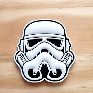 May include: A white and black Stormtrooper helmet design. The helmet features a distinctive shape with black eye slits and a detailed respirator. The design is set against a light brown wooden background.