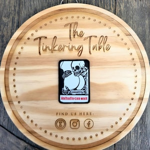 Puede incluir: Un letrero redondo de madera con la inscripci&oacute;n "The Tinkering Table". Una pegatina en blanco y negro con un dise&ntilde;o de calavera y el texto "Valhalla can wait" est&aacute; adherida. Se ven iconos de redes sociales.