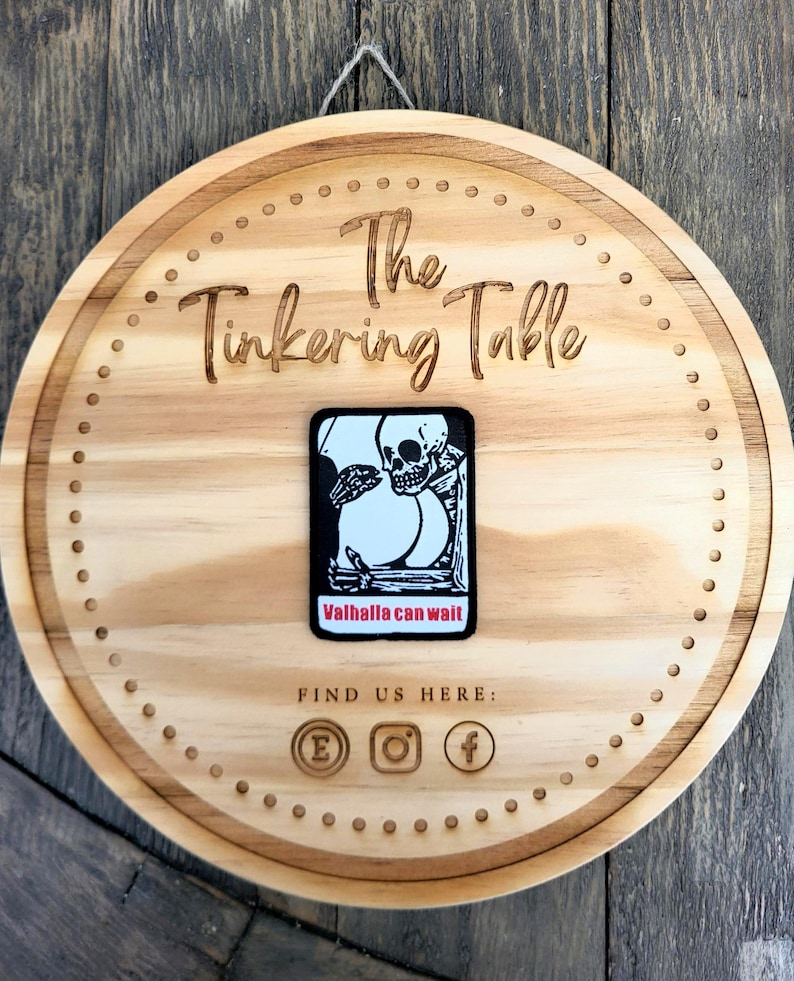 Puede incluir: Un letrero redondo de madera con la inscripci&oacute;n "The Tinkering Table". Una pegatina en blanco y negro con un dise&ntilde;o de calavera y el texto "Valhalla can wait" est&aacute; adherida. Se ven iconos de redes sociales.
