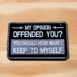 May include: Black rectangular patch with white text that reads "MY OPINION OFFENDED YOU? YOU SHOULD HEAR WHAT I KEEP TO MYSELF." The patch has a black border and is set against a light wood grain background.