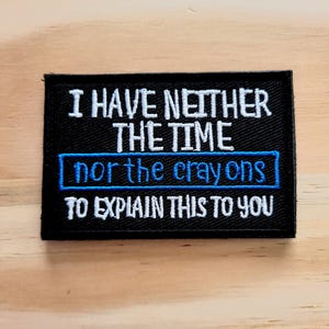 May include: Black rectangular patch with white embroidered text: "I HAVE NEITHER THE TIME nor the crayons TO EXPLAIN THIS TO YOU." The words "nor the crayons" are in a blue box. The patch is on a light wood surface.