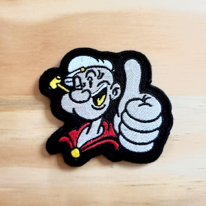May include: Embroidered patch featuring the cartoon character Popeye giving a thumbs-up. The patch has a black border, with Popeye in gray, white, red, and yellow. The design is a classic cartoon style.