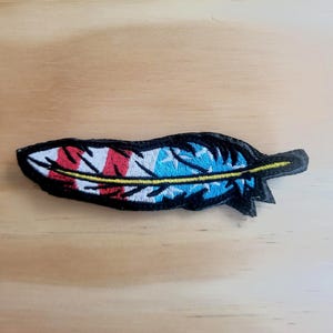 May include: Embroidered feather patch with a patriotic design. The patch features red and white stripes, blue stars, and a yellow accent, all outlined in black. The patch is approximately 4 inches long.
