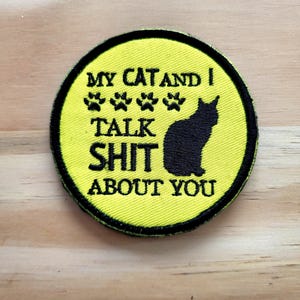 May include: A round, yellow patch with black text and a black cat silhouette. The text reads "MY CAT AND I TALK SHIT ABOUT YOU" with paw prints. The patch has a black border.