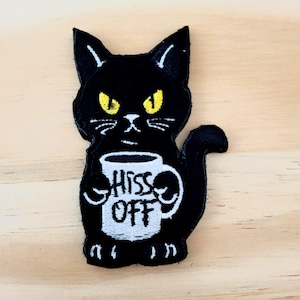 May include: Black cat patch with yellow eyes and the words "Hiss Off" on a white mug. The cat is holding the mug with both paws. The patch is made of black fabric with white and yellow embroidery.