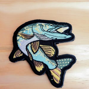 May include: Embroidered patch featuring a detailed depiction of a fish, primarily in shades of blue, yellow, and black. The fish is shown with its mouth open, showcasing intricate stitching and a textured appearance. The patch has a black border.