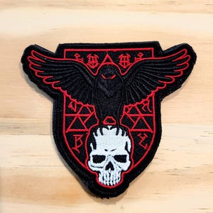 May include: An embroidered patch featuring a black raven with spread wings perched atop a white human skull. The design is outlined in red thread against a black background, incorporating geometric shapes and stylized symbols within the shield shape.