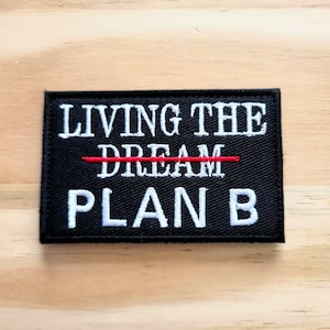 May include: Black rectangular patch with white embroidered text that reads "LIVING THE DREAM" with a red line through "DREAM" and "PLAN B" below. The patch is on a light brown wooden surface.