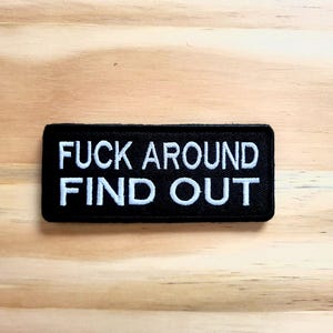May include: Black rectangular patch with white embroidered text that reads "FUCK AROUND FIND OUT". The patch is on a light brown wooden surface.