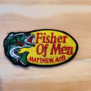 May include: An oval-shaped embroidered patch featuring a green and white fish on the left side and the red text "Fisher Of Men" with "MATTHEW 4:19" underneath on a yellow background, all enclosed by a thick black border.