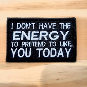 May include: A rectangular black embroidered patch with white stitched text that reads I DON'T HAVE THE ENERGY TO PRETEND TO LIKE YOU TODAY. The patch features a clean border and bold, capitalized lettering in a sans-serif font style.