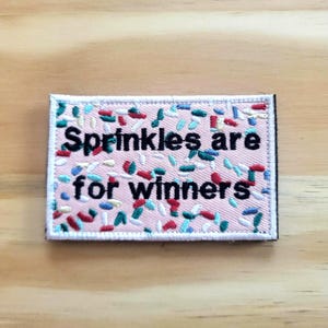 May include: Embroidered patch with the text "Sprinkles are for winners" in black on a pink background. The patch is decorated with colorful sprinkles in red, blue, and green.