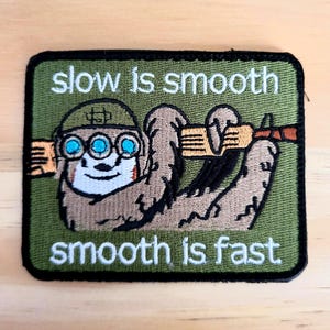 May include: Embroidered patch with a sloth wearing a helmet and goggles, holding a rifle. The patch is rectangular with a green background and black border. Text reads "slow is smooth, smooth is fast."