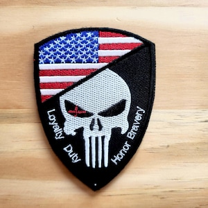 May include: A shield-shaped patch featuring the American flag and a skull design. The top half displays the flag's stars and stripes, while the bottom half shows a white skull on a black background. The words "Loyalty Duty Honor Bravery" are written around the skull.