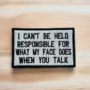 May include: A rectangular patch with the text "I CAN'T BE HELD RESPONSIBLE FOR WHAT MY FACE DOES WHEN YOU TALK" in black on a white background. The patch has a black border and is set against a light wood grain surface.