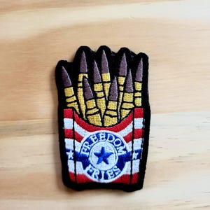 May include: Embroidered patch featuring a design of bullets resembling french fries in a container with red and white stripes. The patch has the words "FREEDOM FRIES" in a circular emblem with a blue star.