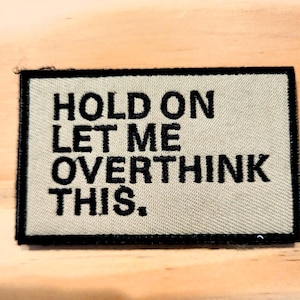 May include: A rectangular patch with the embroidered text "HOLD ON LET ME OVERTHINK THIS." The patch has a tan background with black lettering and a black border. The patch is on a light brown wooden surface.