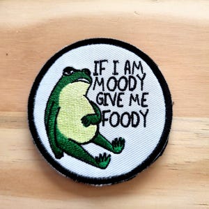 May include: Embroidered patch featuring a green frog with a grumpy expression. The frog is sitting with its belly out. Text on the patch reads "IF I AM MOODY GIVE ME FOODY" in black letters on a white background, surrounded by a black border.