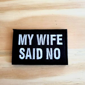 May include: A black rectangular patch with white embroidered text that reads "MY WIFE SAID NO". The patch is set against a light brown wooden surface.