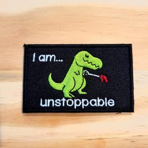 May include: A rectangular black embroidered patch featuring a green Tyrannosaurus rex holding a silver wrench with a red handle. The text "I am..." is embroidered in white above the dinosaur, and the word "unstoppable" is in white below it.
