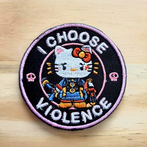 May include: Round embroidered patch featuring a cartoon cat character in a blue and orange outfit, with the text "I CHOOSE VIOLENCE" in white. The patch has a black background with a pink border and small skull icons.