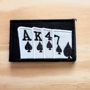 May include: A rectangular black patch featuring a poker hand of spades: Ace, King, 4, and 7. The card faces are white with black spade symbols and text. The patch is likely for clothing or accessories.