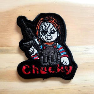 May include: Embroidered patch featuring Chucky, the killer doll, holding a gun. The patch has a black border and the name "Chucky" in red. Chucky is depicted with red hair, a scarred face, and wearing a striped shirt.