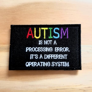 May include: A black embroidered patch with the word "AUTISM" in rainbow colors. The text below reads "IS NOT A PROCESSING ERROR. IT'S A DIFFERENT OPERATING SYSTEM."