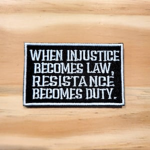 May include: A rectangular black patch with white embroidered text: "WHEN INJUSTICE BECOMES LAW, RESISTANCE BECOMES DUTY." The patch has a white border and is set against a light wood grain background.